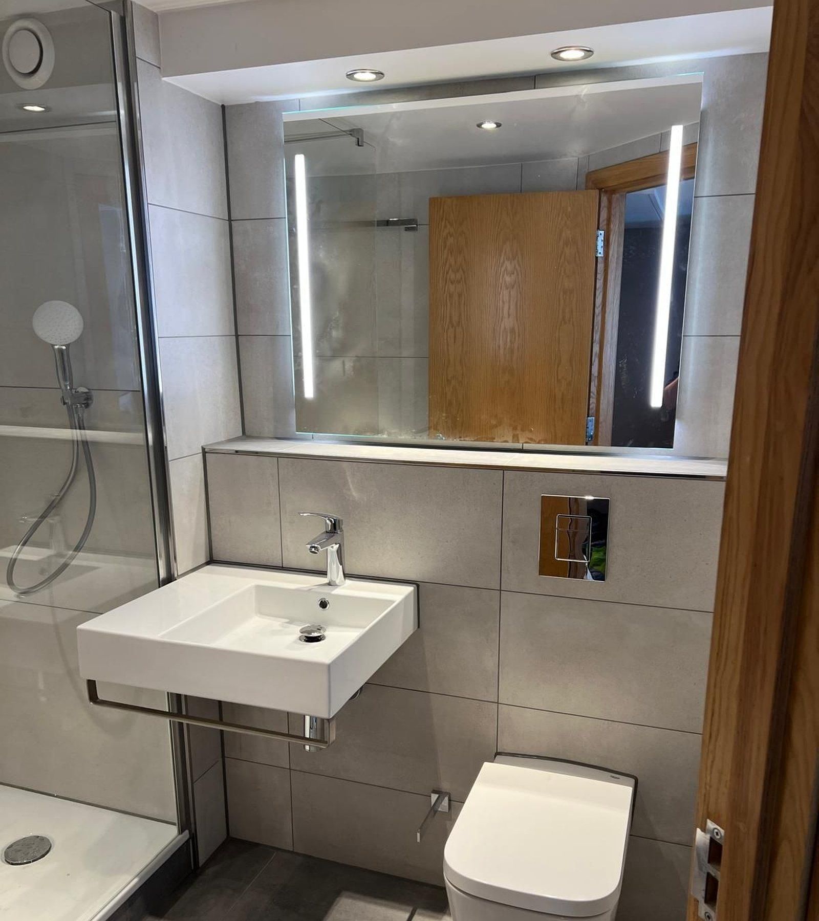 Refurbished hotel guest bathroom with large-format grey tiling, wall-hung basin and backlit mirror